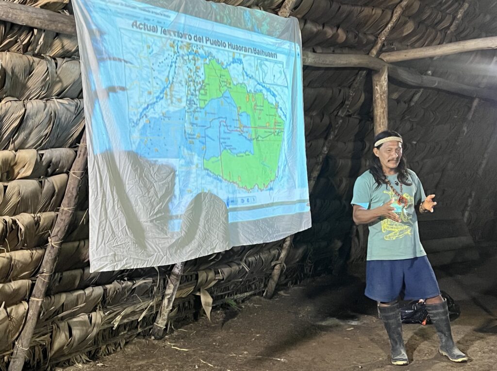 Penti Baihua speaks at a gathering of recently-contacted Waorani people in Gemenoweno, Ecuador beginning on March 11, 2023. He stands before a map depicting the territorial complexities of the Ecuador Amazon region. Credit: Katie Surma/Inside Climate News